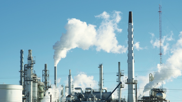 “Refinery pollution; oil refinery smokestacks and machinery against a blue sky”